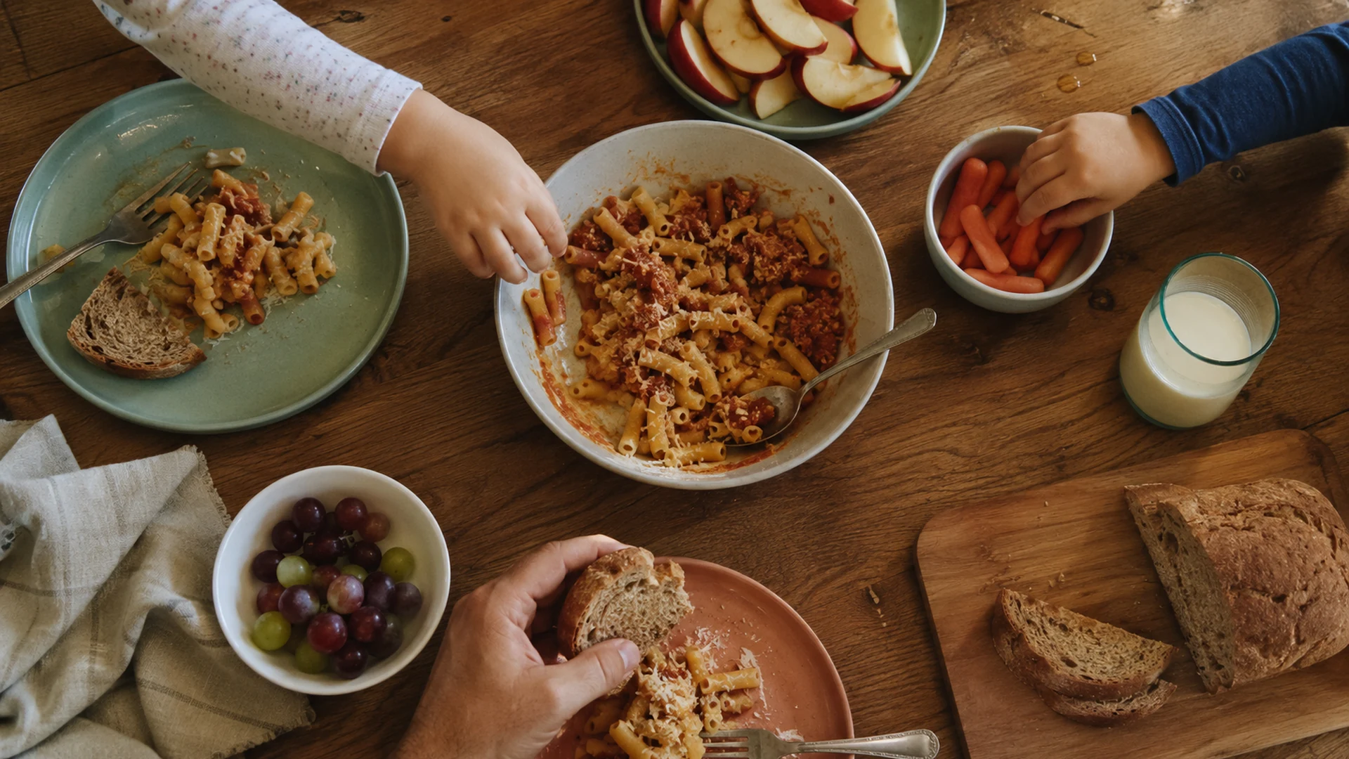 Overhead view of an adult and two children sharing pasta, fruit, carrots, bread, and milk at a wooden dinner table