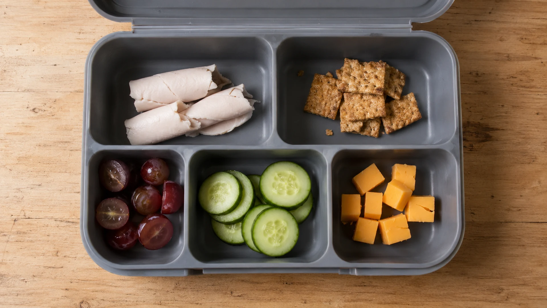 An open gray divided lunchbox with turkey roll-ups, crackers, grapes, cucumber rounds, and cheese cubes on a wooden table